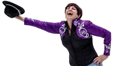 A landscape photo of Gail Taylor dressed in a black and purple top, with her right arm stretched out holding a hat.