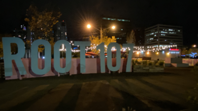 A photo of the baby blue Root 107 sign at night, with lights visible in the background.