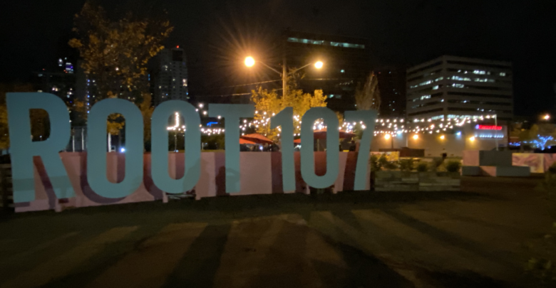 A photo of the baby blue Root 107 sign at night, with lights visible in the background.