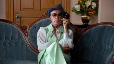 A screenshot of a scene in "Swan Song" where Pat sits on a couch with his legs crossed.