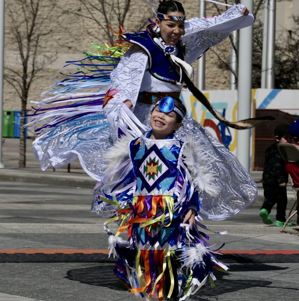 A child smiles while dressed in regalia with an older dancer for Moose Hide Campaign Day