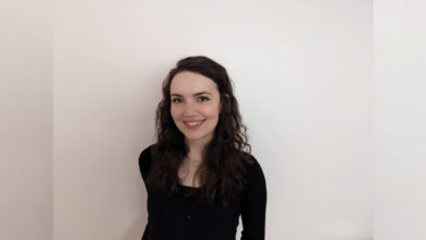 Ally McIntyre stands in the middle of a white wall in a black blouse