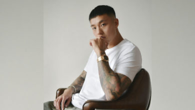 Anders sits in a brown chair with a white t-shirt on. His left arm is up to his face.